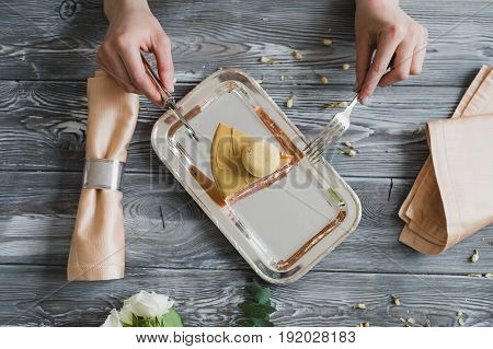Top view of hands going to eat dessert, yellow mousse cake with almond dacquoise, raspberry confit, crispy layer with caramelized hazelnuts and raspberry powder, mousse based on bitter chocolate served on wooden table.