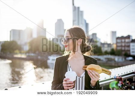 Young businesswoman having a snack with traditional Franfurt sausage and coffee on the bridge with skyscrapers on the background in Frankfurt during the sunset