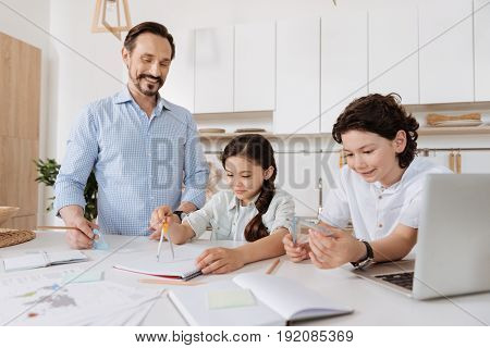 Exciting novelties. Cheerful handsome boy holding a set square while his sister using a pair of compasses and their pleasant young father looking at them with an affectionate smile