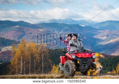 Young Couple On Red Quad Bike Ride In Mountains. Happy Man Driving The Atv, Girlfriend Pointing At S