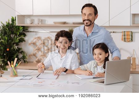 Happy together. Charming young father hugging his lovely children from behind while his son using a compass and his daughter making notes and all of them looking at the camera with sincere smiles