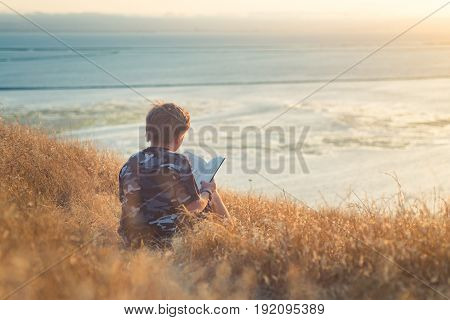 Boy reading book in yellow grass high in the hills, watching beautiful sunset above Bay Area, rear view