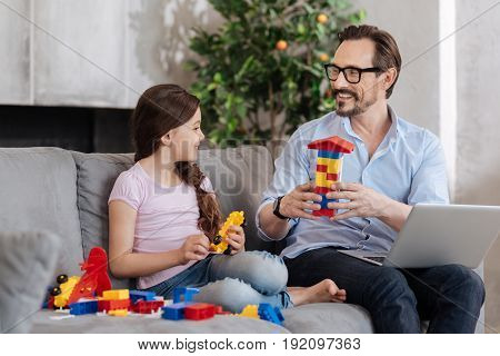 Pleasant discussion. Petite gorgeous girl and her pleasant young father sitting on the sofa and discussing their new erector set while holding some pieces of it each