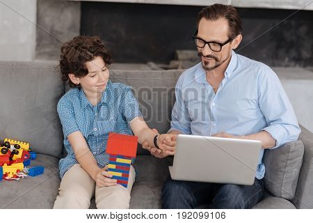 Happy together. Pleasant young father sitting on the sofa with his laptop on the lap and touching the hand of his son holding a tower built with an erector set