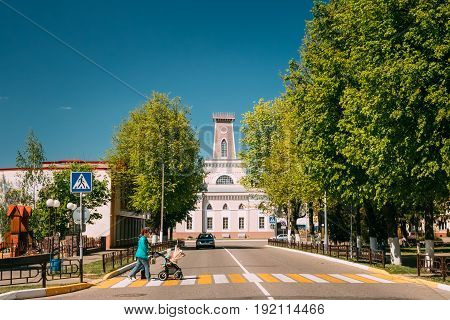 Chachersk, Belarus - May 14, 2017: Woman With A Stroller Crosses The Road On A Crossing. Old City Hall In Sunny Summer Day On Background. Pedestrian Town Hall In Chechersk