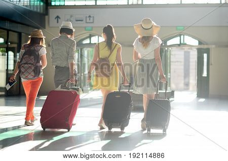 Group of young tourists at the station. In hands at girls suitcases tickets passports. Summer tourism. View from a back.