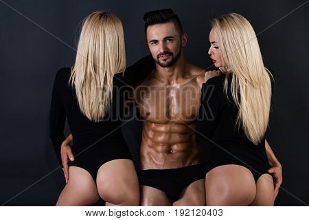 love relations. athletic man with muscular wet body and strong torso with sexy blonde girls in underwear pants and bodysuit on black background lesbian and gay homosexual seduction and foreplay