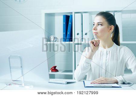 Pensive young business woman sitting at the dask with desktop monitor and looking at the window.