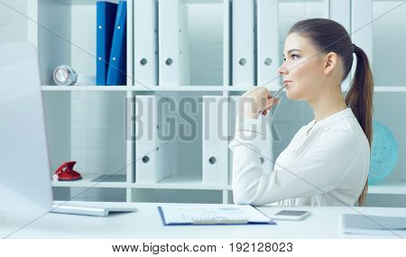 Pensive young business woman sitting turning sideways at the dask with desktop monitor and looking at the window.