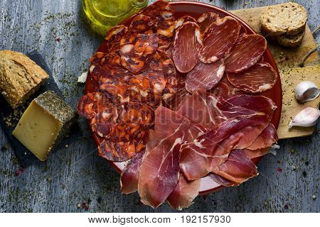 high-angle shot of a plate with an assortment of different spanish cold meats as chorizo, cured pork tenderloin and serrano ham, a piece of manchego cheese, bread and a cruet with olive oil on a table
