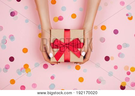 Woman hands holding present box with red bow on pastel pink background with multicolored confetti. Flat lay style.