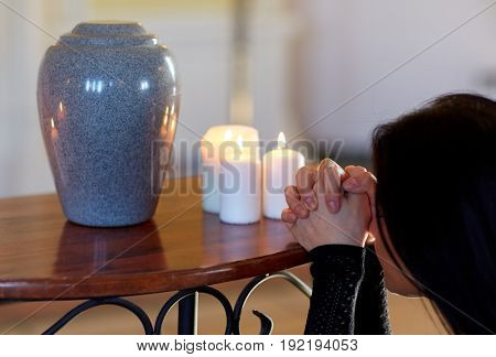 cremation, people and mourning concept - sad woman with cinerary urn and candles praying at funeral in church
