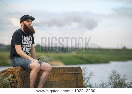 Vape Bearded Man In Real Life. Portrait Of Young Guy With Large Beard In A Cap And Sunglasses Having