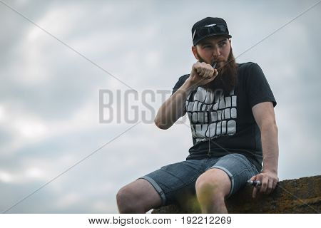 Vape Bearded Man In Real Life. Portrait Of Young Guy With Large Beard In A Cap And Sunglasses Having