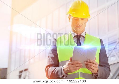 Young male engineer in reflector-vest and hardhat using digital tablet outside industry