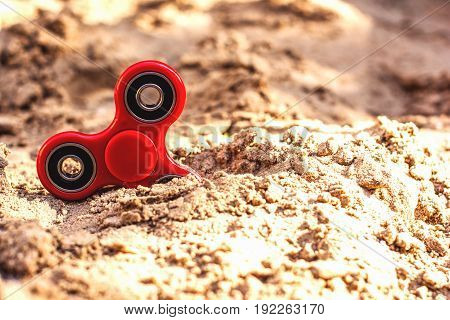 Popular spinner gadget in 2017 against the background of sand. Modern red plastic spinner fidget game for young people. Learn spinning tricksimprove concentration.