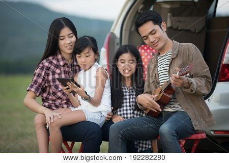 Happy little girl playing ukulele with asian family sitting in the car for enjoying road trip and summer vacation in camper van