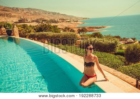 Caucasian sexy skinny model with thin waist relaxing in luxurious infinity pool. Young woman rest in spa resort. Summer luxury vacation. Tourism and travel concept. Enjoying suntan. Dead Sea, Jordan.