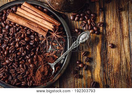 Coffee Beans with Spooonful of Ground Coffee Cinnamon Sticks and Chinese Star Anise on Metal Plate. Some Beans Scattered on Wooden Table. View from Above.