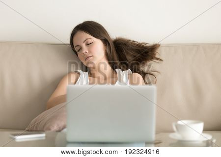 Young attractive woman falling asleep on sofa at home in front of laptop. Teenager girl resting after long hours surfing Internet. Female student takes break after finishing computer work, exam study