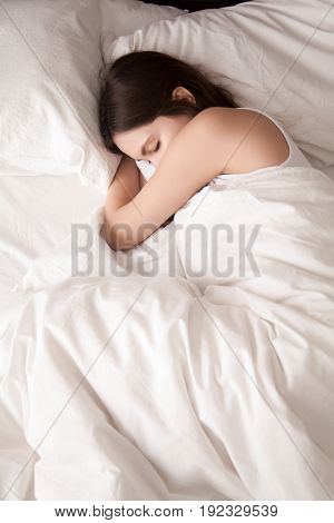 Tired young woman sleeping well on the side in bed with white sheets, resting after sleepless night. Female student lying in bed until late morning. Getting enough sleep concept. View from above
