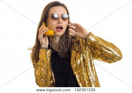 funny woman in round sunglasses waers a golden jacket and talking in banana like a mobile phone isolated on white background