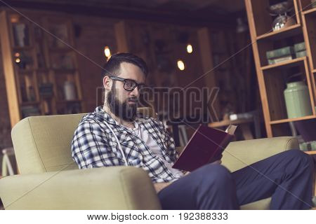 Young man sitting in a cozy armchair in a cafe reading a book and enjoying his leisure time