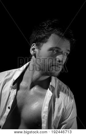 Black and white portrait of young, attractive man in unbottoned, creased white shirt sitting in the dark