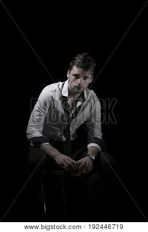 Portrait of young, attractive man in unbottoned, creased white shirt with upturned sleeves and loose tie sitting in the dark