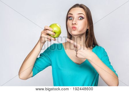 Young beautiful woman with freckles and green dress holding and eating apple and pointing and looking at camera with big eyes. studio shot isolated on light gray background.