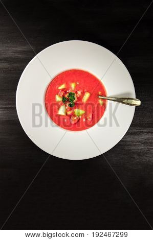 An overhead photo of a plate of gazpacho soup with slices of tomato, cucumber, and onion, with parsley leaves, on a dark background with a place for text