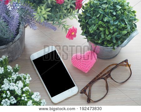 wood table with blank screen for text on smartphone tablet cell phone pink heart shape sign modern eyeglasses and beautiful flower on pot view from top wood table.