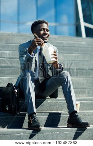 Happy businessman with chopsticks eating food brom box on staircase