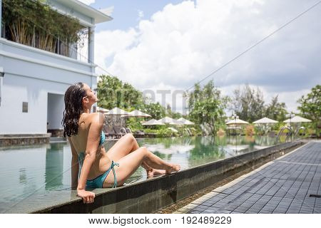 Relaxed young Caucasian woman wearing bikini sitting on pool edge and sunbathing at hotel during summer vacations
