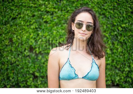 Portrait of smiling young Caucasian woman wearing sunglasses and bikini posing at bush