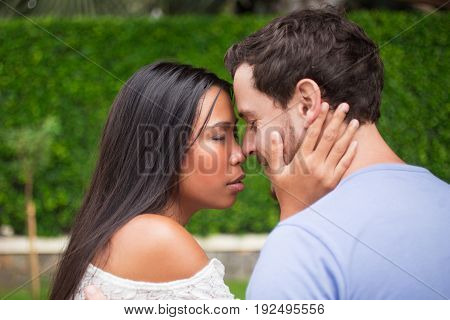 Closeup of affectionate young interracial couple touching noses and hugging with their eyes closed and blurred green leaves wall in background. Woman embracing boyfriend neck. Side view.