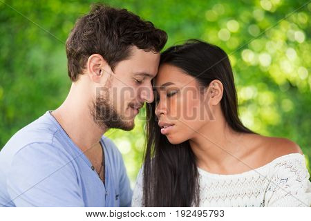 Closeup of affectionate young interracial couple touching foreheads with their eyes closed and blurred green leaves wall in background