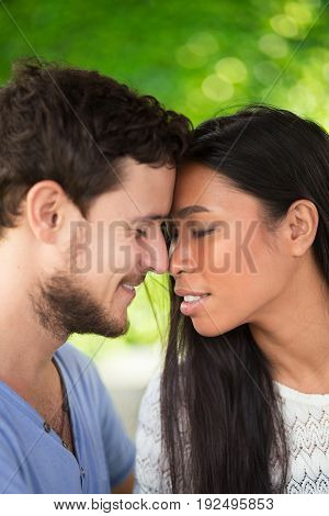 Closeup of affectionate young interracial couple touching foreheads with their eyes closed and blurred green leaves wall in background