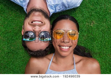 Closeup portrait of smiling young multi-ethnic couple looking at camera and lying on lawn. Top view.