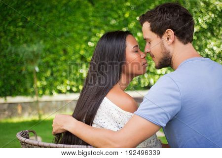 Closeup of affectionate young interracial couple touching noses and hugging with their eyes closed and blurred green leaves wall in background. Side view.