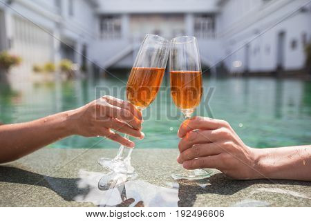 Cropped view of young couple clinking glasses of champagne at edge of swimming pool with building in background. Side view.