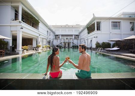 Smiling young interracial couple looking at each other, clinking glasses of champagne and sitting at edge of swimming pool with building in background. Back view.