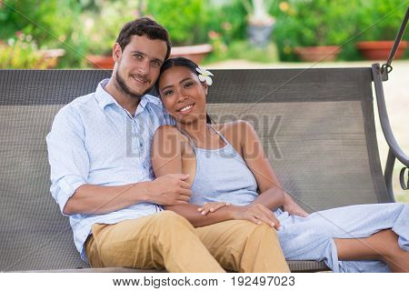 Closeup portrait of smiling young Caucasian man embracing Asian girlfriend. They are looking at camera and sitting on swinging bench with blurred plants in background.