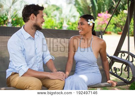 Closeup portrait of smiling young Caucasian man and his Asian girlfriend looking at each other and sitting on swinging bench with blurred plants in background