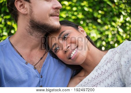 Closeup of smiling young Asian woman leaning head on Caucasian boyfriend shoulder with green leaves wall in background. Cropped view.