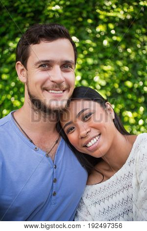 Closeup of smiling young Asian woman leaning head on Caucasian boyfriend shoulder with green leaves wall in background. They are looking at camera.