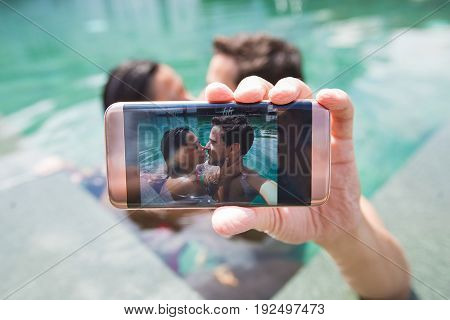 Closeup of content young interracial couple taking selfie photo at edge of swimming pool with focus on smartphone screen. Front view.