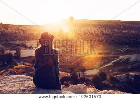A tourist girl in a hat sits on a mountain and looks at the sunrise and balloons in Cappadocia. Tourism, Turkey.