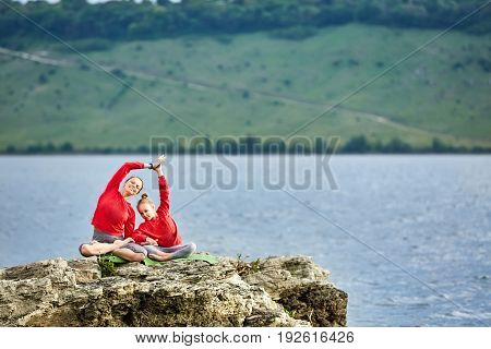 Sporty mother and daughter doing yoga on the rock near beautiful river. Beautiful mother and daughter dressed in the sportwear and barefoot. Horizontal photo with beauty landscape. Concept of the healthy and active lifestyle.
