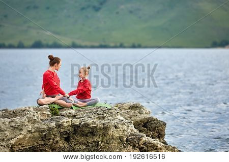 Sporty mother and daughter doing yoga on the rock near beautiful river. Beautiful mother and daughter dressed in the sportwear and barefoot. Horizontal photo with beauty landscape. Concept of the healthy and active lifestyle.